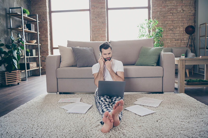 02 Un hombre con barba sentado en la alfombra con una laptop y hablando por telefono