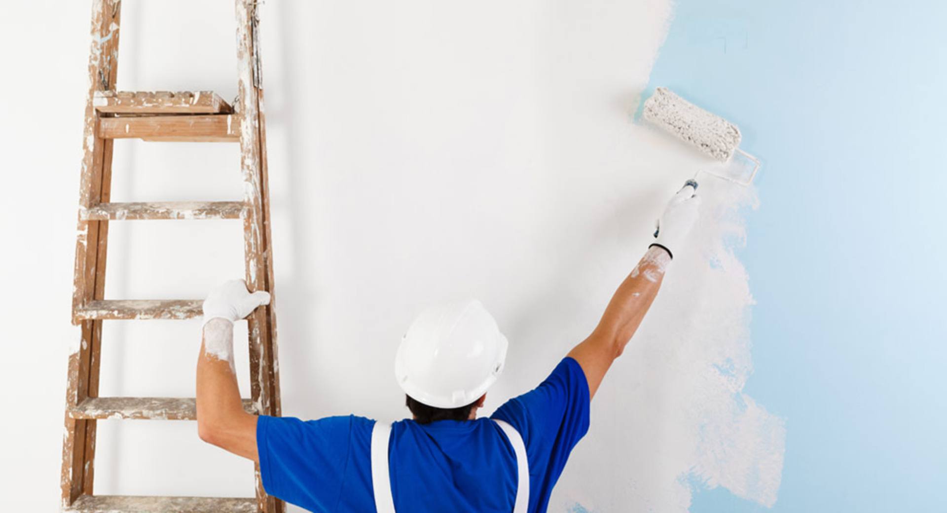 Hombre con casco aplicando pintura impermeabilizante blanca a una pared usando un rodillo desde una escalera